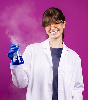 Madeleine holding a beaker of blue liquid while wearing a lab coat and protective glasses.
